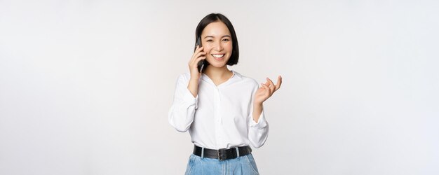 Glimlachende gelukkige aziatische vrouw die op smartphone praat met de verkoopster van de klant die mobiele telefoon vasthoudt en gebaren maakt over een witte achtergrond
