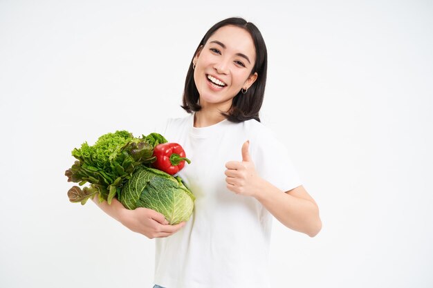 Glimlachende gelukkige aziatische vrouw toont duimen omhoog en groenten die gezond ruw voedsel eten dat over wit wordt geïsoleerd