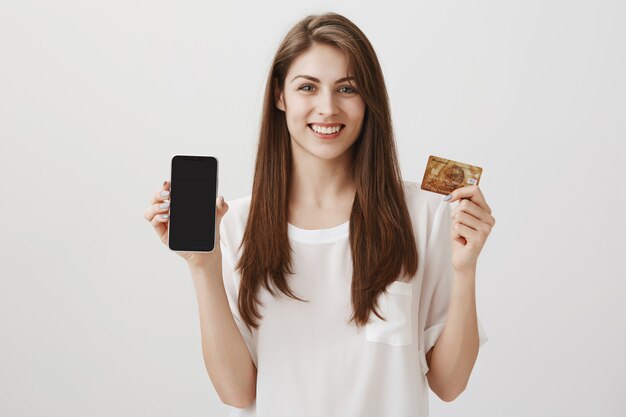 Glimlachende gelukkige vrouw die mobiele telefoonvertoning en creditcard toont. Promo van winkelapplicatie