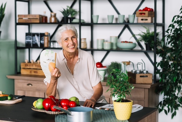 Gratis foto glimlachende hogere de ventilator in hand zitting van de vrouwenholding achter keukentop