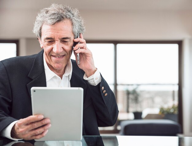 Glimlachende hogere mens die op mobiele telefoon spreekt die digitale tablet op het werk bekijkt