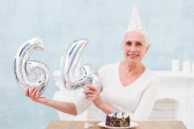 Gratis foto glimlachende hogere vrouw die de ballon van de 64 aantalfolie met haar verjaardagscake op lijst tonen