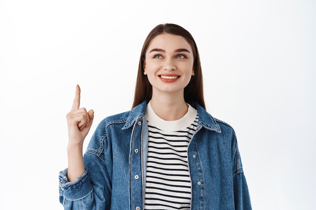 Glimlachende hoopvolle studente, wijzend en opzoekend met een doordachte glimlach, promotionele tekst naar boven lezend, taalcursussen of productadvertentie, staande op een witte achtergrond