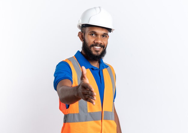 Glimlachende jonge Afro-Amerikaanse bouwer man in uniform met veiligheidshelm stak zijn hand uit geïsoleerd op een witte achtergrond met kopie ruimte