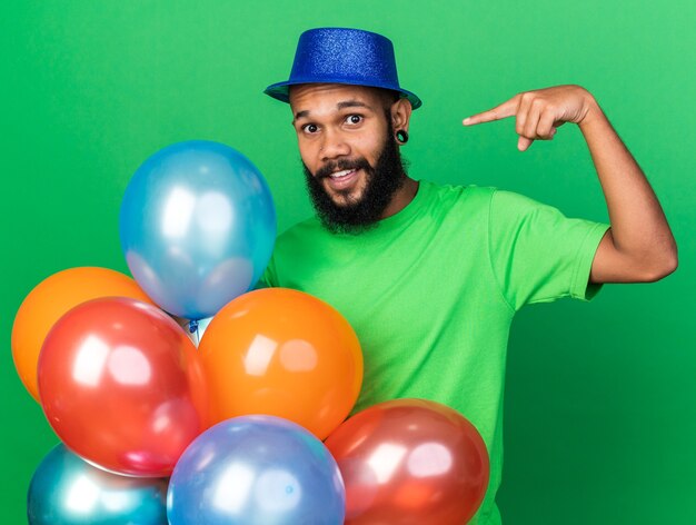 Glimlachende jonge Afro-Amerikaanse man die een feestmuts draagt en naar ballonnen wijst