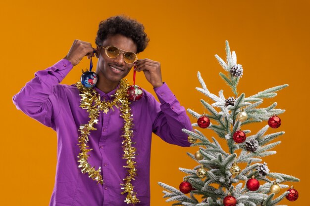 Glimlachende jonge Afro-Amerikaanse man met bril met klatergoud slinger rond de nek staande in de buurt van versierde kerstboom opknoping kerstballen op oren geïsoleerd op oranje muur