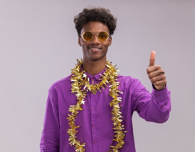 Glimlachende jonge Afro-Amerikaanse man met bril met klatergoud slinger rond nek kijken camera weergegeven: duim omhoog geïsoleerd op een witte achtergrond