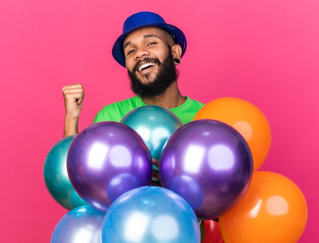 Glimlachende jonge Afro-Amerikaanse man met een feestmuts die achter ballonnen staat en een ja-gebaar toont dat op een roze muur is geïsoleerd