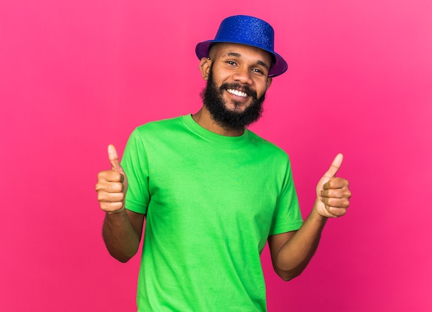 Glimlachende jonge Afro-Amerikaanse man met een feestmuts die duimen laat zien die op roze muur is geïsoleerd