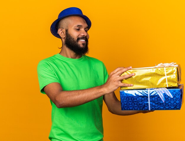 Glimlachende jonge Afro-Amerikaanse man met een feestmuts die geschenkdozen vasthoudt en bekijkt