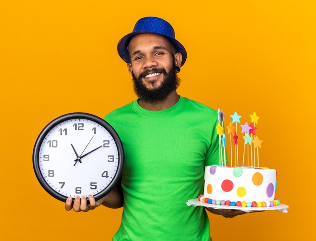 Glimlachende jonge Afro-Amerikaanse man met een feestmuts met cake met wandklok