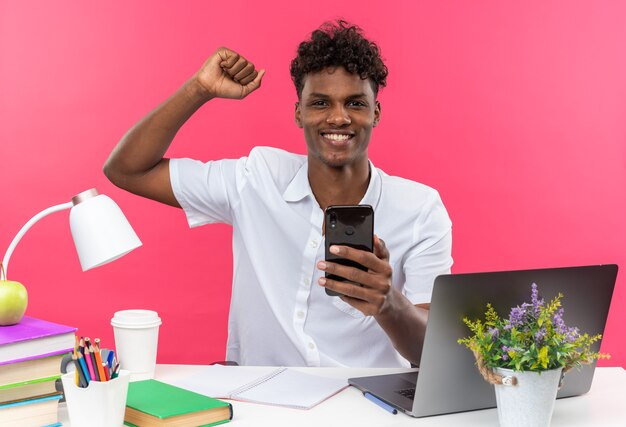 Glimlachende jonge Afro-Amerikaanse student die aan een bureau zit met schoolhulpmiddelen die de telefoon vasthouden en de vuist omhoog houden