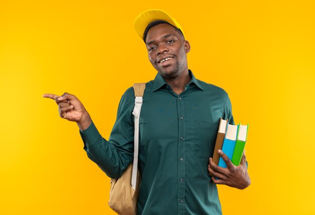 Glimlachende jonge Afro-Amerikaanse student met pet en rugzak die boeken vasthoudt en naar de zijkant wijst, geïsoleerd op een oranje muur met kopieerruimte