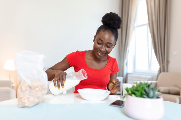 Glimlachende jonge Afro-Amerikaanse vrouw giet cornflakes in bord met melk Het meisje heeft 's ochtends een gezond ontbijt op een stijlvol, gezellig huis terwijl ze haar e-mail controleert op laptop