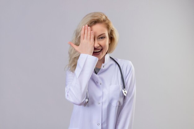 Glimlachende jonge arts die stethoscoop in medische toga draagt bedekt oog op witte muur