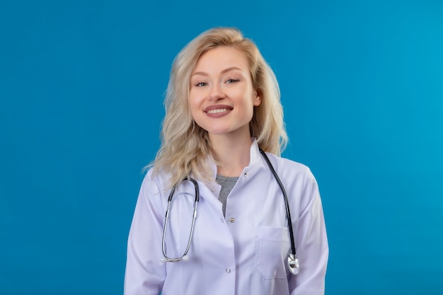 Glimlachende jonge arts die stethoscoop in medische toga draagt op blauwe muur
