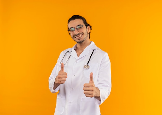 Glimlachende jonge arts met medische bril die medische mantel met stethoscoop draagt zijn duimen omhoog op geïsoleerde gele muur met exemplaarruimte