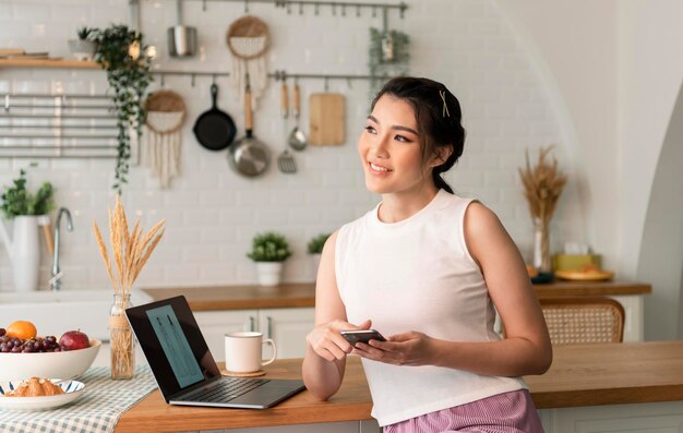 Glimlachende jonge aziatische vrouw die mobiele telefoon gebruikt terwijl ze thuis met laptop in de keuken zit