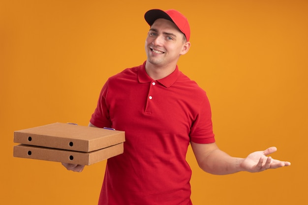 Glimlachende jonge bezorger die eenvormig met GLB draagt die pizzadozen houdt die hand spreidt die op oranje muur wordt geïsoleerd