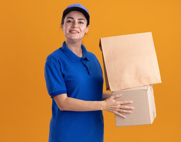 Glimlachende jonge bezorger in uniform en pet met kartonnen doos met papieren pakket erop geïsoleerd op oranje muur