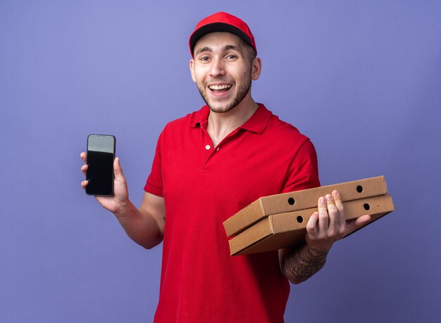 Glimlachende jonge bezorger in uniform met pet die pizzadozen vasthoudt met telefoon