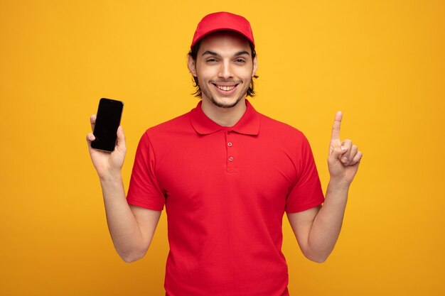 Glimlachende jonge bezorger met uniform en pet kijkend naar camera met mobiele telefoon die omhoog wijst geïsoleerd op gele achtergrond