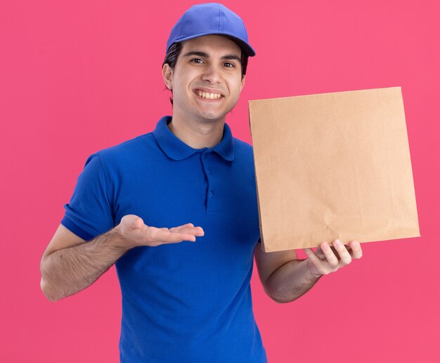 Glimlachende jonge blanke bezorger in blauw uniform en pet die vasthoudt en met de hand naar een papieren pakket wijst