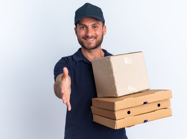 Glimlachende jonge blanke bezorger in blauw uniform en pet met pizzapakketten en kartonnen doos kijkend naar camera die begroetingsgebaar doet geïsoleerd op een witte muur met kopieerruimte