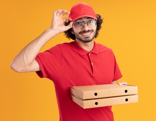 glimlachende jonge blanke bezorger in rood uniform en pet met een bril die een pet vasthoudt met pizzapakketten geïsoleerd op een oranje muur orange