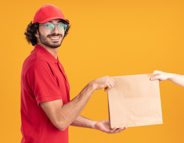 Glimlachende jonge blanke bezorger in rood uniform en pet met een bril in profielweergave die een papieren pakket aan de klant geeft