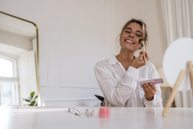 Glimlachende jonge blanke dame gebruikt blozen in het dagelijks leven terwijl ze binnenshuis aan tafel zit. Cosmetische procedures concept