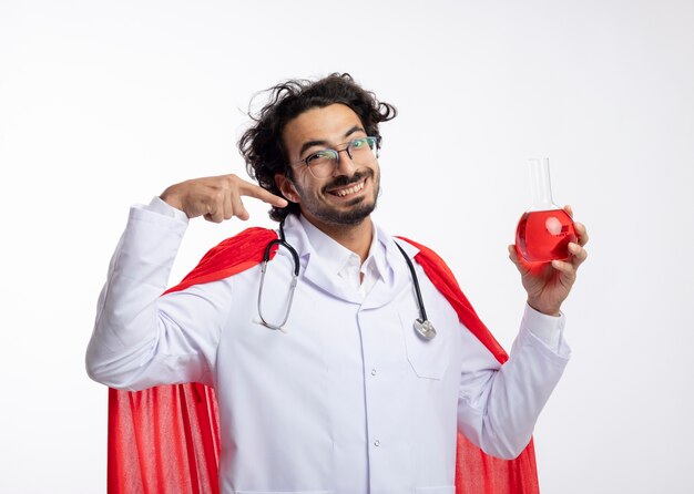 Glimlachende jonge blanke man in optische bril dragen arts uniform met rode mantel en met een stethoscoop om de nek houdt en wijst naar rode chemische vloeistof in glazen kolf op witte muur