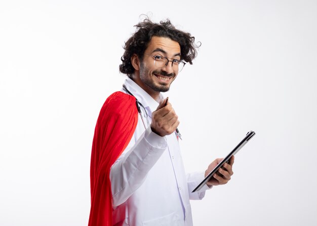 Glimlachende jonge blanke man in optische bril dragen arts uniform met rode mantel en met een stethoscoop om de nek staat zijwaarts met klembord en wijst met potlood