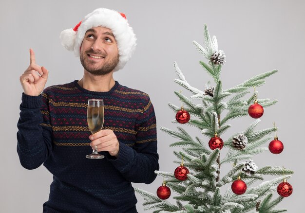 Glimlachende jonge blanke man met kerstmuts staande in de buurt van de kerstboom met glas champagne op zoek en omhoog geïsoleerd op een witte achtergrond