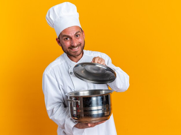 Glimlachende jonge blanke mannelijke kok in uniform van de chef en dop die deksel van de pot opent opening