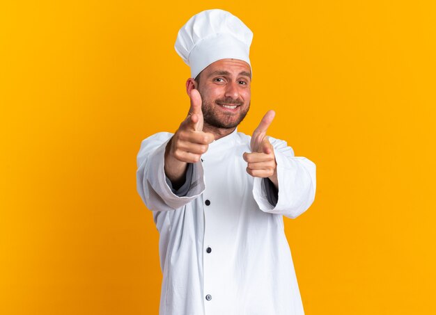 Glimlachende jonge blanke mannelijke kok in uniform van de chef-kok en pet die een pistoolgebaar doet