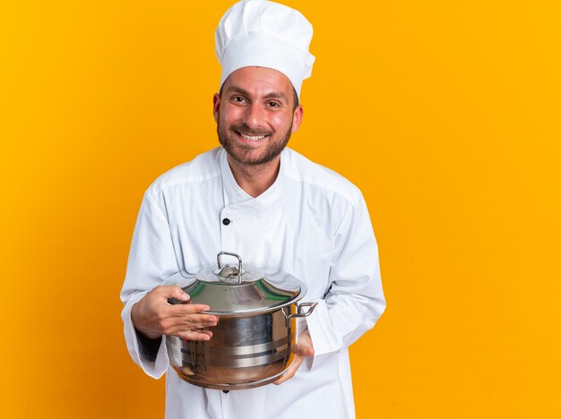 Glimlachende jonge blanke mannelijke kok in uniform van de chef-kok en pet met pot kijkend naar camera geïsoleerd op oranje muur met kopieerruimte