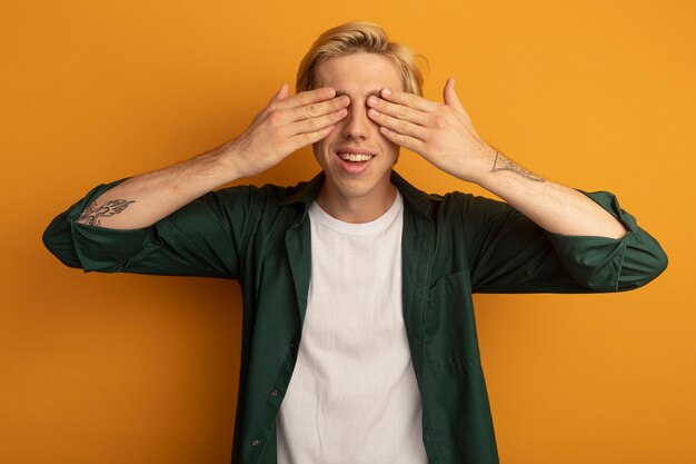 Glimlachende jonge blonde kerel die groene t-shirt draagt ?? bedekte ogen met handen