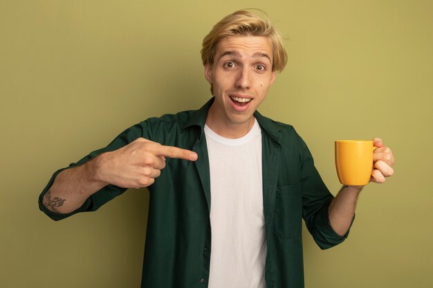 Glimlachende jonge blonde kerel die groene t-shirtholding draagt ?? en wijst op kopje thee