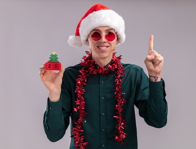 Glimlachende jonge blonde man met kerstmuts en bril met klatergoud slinger om nek met kerstboom speelgoed met datum kijken camera omhoog geïsoleerd op witte achtergrond