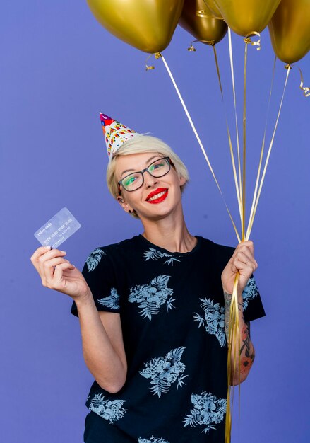 Glimlachende jonge blonde partijvrouw die glazen en verjaardag GLB houden die ballons en creditcard houden die voorzijde bekijken die op purpere muur wordt geïsoleerd