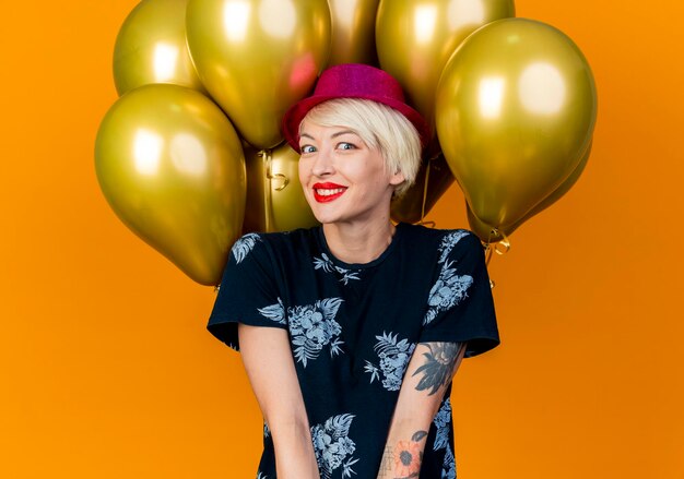Glimlachende jonge blonde partijvrouw die partijhoed draagt die zich voor ballons bevindt die voorzijde bekijken die op oranje muur wordt geïsoleerd