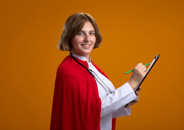 Glimlachende jonge blonde superheldvrouw in rode cape, doktersuniform en stethoscoop dragen, staande in profielweergave schrijven met potlood op klembord, geïsoleerd