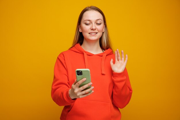 Glimlachende jonge blonde vrouw die en mobiele telefoon bekijkt die ernaar zwaait