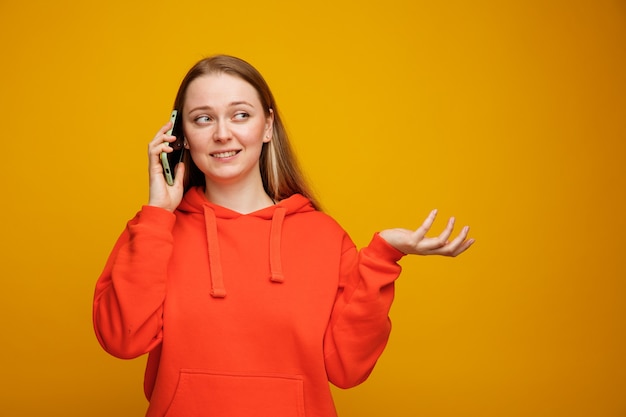 Glimlachende jonge blonde vrouw die op telefoon spreekt die kant bekijkt die lege hand toont