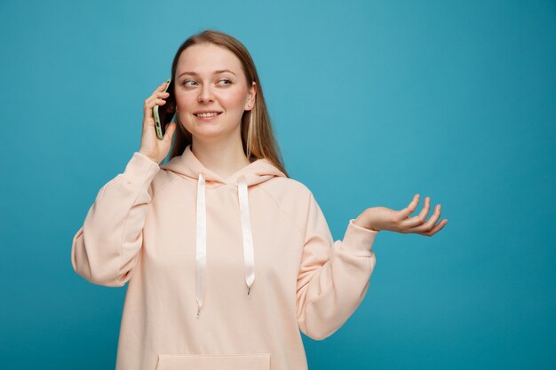 Glimlachende jonge blonde vrouw die op telefoon spreekt die kant bekijkt die lege hand toont