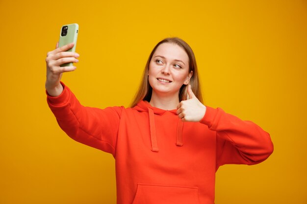 Glimlachende jonge blonde vrouw die selfie neemt die duim toont