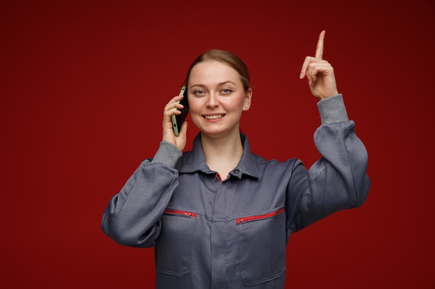 Glimlachende jonge blonde vrouwelijke ingenieur die uniform draagt ?? die op telefoon spreekt die omhoog wijst