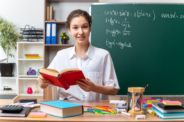 Glimlachende jonge blonde vrouwelijke wiskundeleraar die aan het bureau zit met schoolhulpmiddelen die een boek in de klas houden
