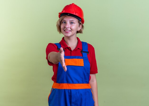 Glimlachende jonge bouwvrouw in uniform die hand uitsteekt aan de voorkant geïsoleerd op olijfgroene muur